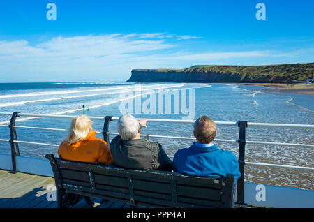A  lady and two men  sitting on Saltburn Pier looking south towards Huntcliff and watching the surfers Stock Photo
