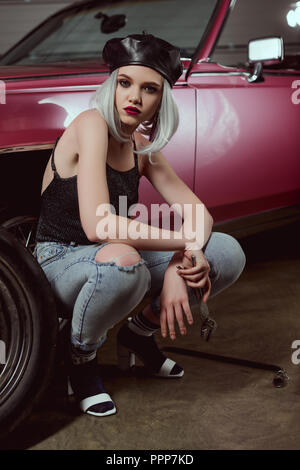 stylish blonde girl in beret holding wrench and looking at camera while repairing car Stock Photo