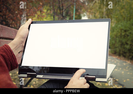 casual man sitting on a bench in the autumn Park with his legs crossed and presenting his laptop with white blank screen. Touch screen on laptop Stock Photo