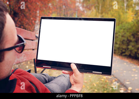 man lies on a bench in the autumn Park and looks at the screen of a modern ultrabook transformer. blank screen for design. Outdoor recreation. Stock Photo