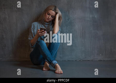 Helpline, psychological assistance. Suffering young woman sitting in a corner with a phone in her hand. mental health, grey background Stock Photo