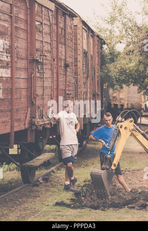 Jaworzyna Slaska, Poland - August 2018 : Two young men standing by an old wooden train carriage looking at digger at work Stock Photo