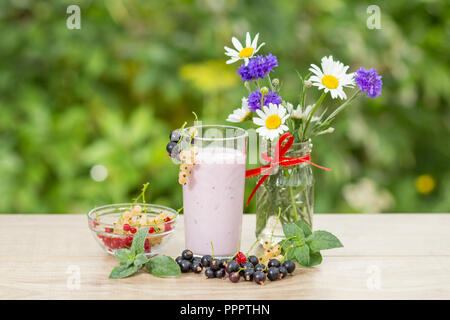 Glass of delicious currant yogurt with fresh red, black and white currants in a bowl and leaves of mint, chamomile and cornflowers in vase on a wooden Stock Photo