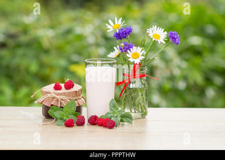 Glass of delicious raspberry yogurt with fresh raspberries, homemade strawberry jam in a jar and leaves of mint, chamomile and cornflowers in vase on  Stock Photo