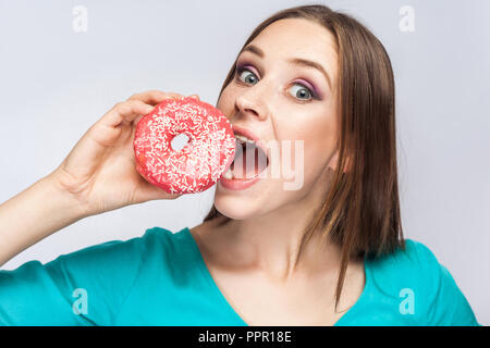 Portrait of hungry young beautiful girl in blue blouse standing, holding and trying to bite big pink donut with open mouth and big eyes in grey backgr