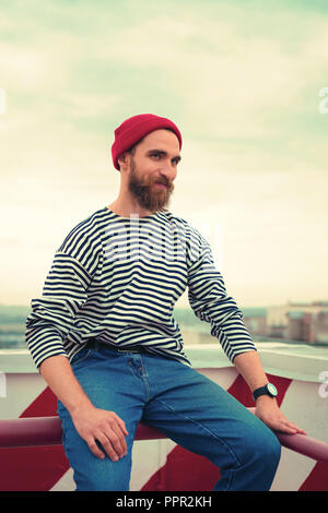 Happy bearded man in red hat smiling while sitting on the roof Stock Photo