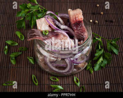 In a glass jar is a salted herring fillet cut into pieces sprinkled with green and red onion and pepper Stock Photo