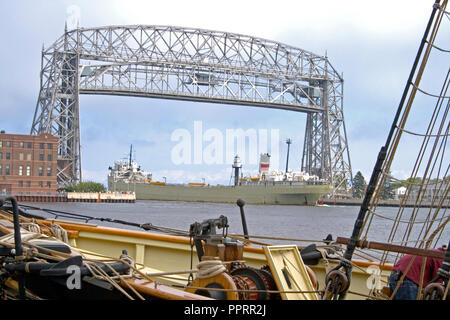 Long cargo ship passing under the Aerial Lift Bridge in the Duluth Port of Lake Superior, Pride of Baltimore Tall Ship below. Duluth Minnesota MN USA Stock Photo