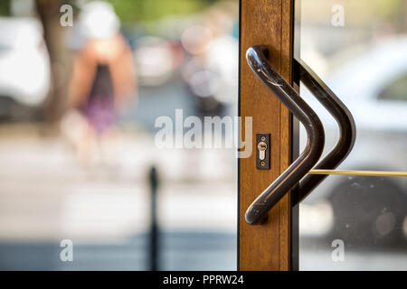 Close-up of plastic or wooden modern brown door with transparent glass surface and big metal handle on blurred bright copy space bokeh background. Ent Stock Photo