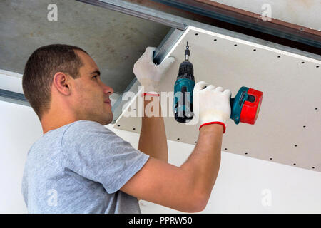 Young man in usual clothing and work gloves fixing drywall suspended ceiling to metal frame using electrical screwdriver on ceiling insulated with shi Stock Photo