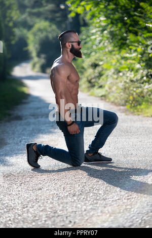 Handsome Young Model Kneeling Strong Outdoors and Flexing Muscles - Muscular Athletic Bodybuilder Man Posing Stock Photo