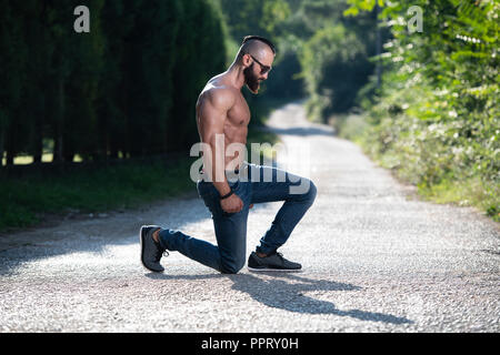 Handsome Young Model Kneeling Strong Outdoors and Flexing Muscles - Muscular Athletic Bodybuilder Man Posing Stock Photo