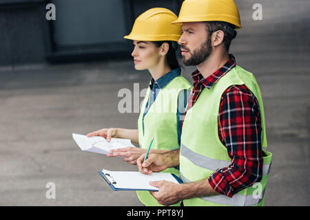 architects in helmets working with blueprint and clipboard on roof Stock Photo