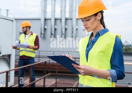 female architect in helmet writing in clipboard on roof, male colleague with blueprint behind Stock Photo