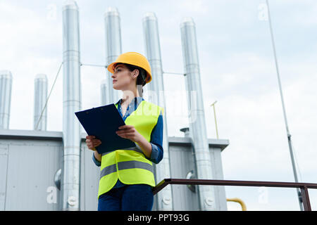 beautiful female builder in safety vest and hardhat writing in clipboard on industrial construction Stock Photo