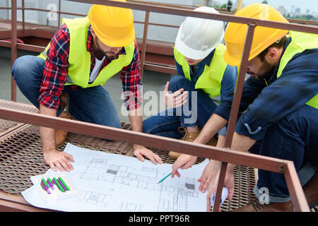 three architects in hardhats working with blueprints on roof Stock Photo