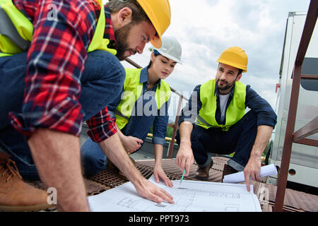 three engineers in hardhats working with blueprints on roof Stock Photo