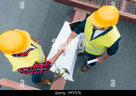 overhead view of engineers in helmets with blueprints shaking hands on roof Stock Photo