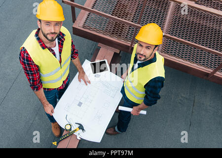 overhead view of male engineers in safety vests and helmets working with blueprints, digital tablet and tools on roof Stock Photo