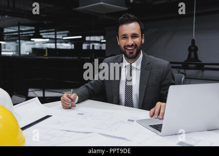 smiling male architect working with blueprints and laptop Stock Photo