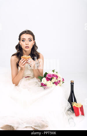 shocked young bride in wedding dress sitting on floor with burger in hand on white Stock Photo