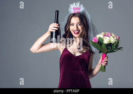 smiling future bride in veil for bachelorette party with bouquet and champagne looking at camera isolated on grey Stock Photo