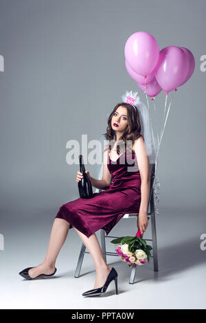 depressed future bride in veil for bachelorette party sitting on chair with champagne and flowers and looking at camera after party on grey Stock Photo