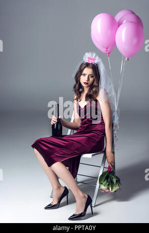 sad future bride in veil for bachelorette party sitting on chair with champagne and flowers and looking at camera after party on grey Stock Photo