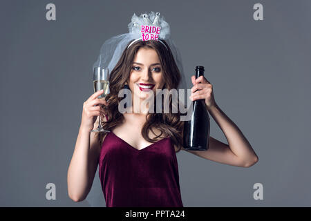 beautiful future bride in veil for bachelorette party with champagne bottle and glass looking at camera isolated on grey Stock Photo