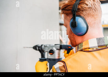 confident builder in noise reducing headphones using power drill at construction site Stock Photo