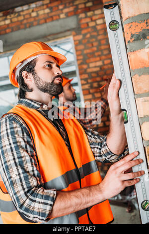 side view of handsome builder using bubble level at construction site while his colleague working on background Stock Photo