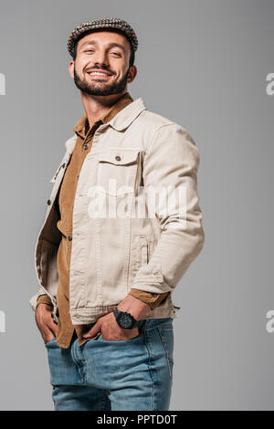 bearded smiling man in autumn jacket and tweed cap posing with hands in pockets, isolated on grey Stock Photo