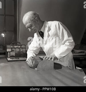 1950s, historical, leatherworking, a male craftsman in a white coat using a sharp scalpel knife and wooden frame to mark a piece of leather before cutting. Stock Photo