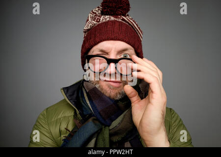 Portrait of a funny mature man in warm winter clothes looking at camera through glasses over gray background. Stock Photo