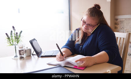 Plus size business woman busy multitasking at her local coffee shop to get as much work done as possible early in the morning, as she writes in her jo Stock Photo