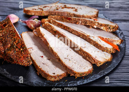Close up on slices of roasted ham coated in spices: paprika, chili flakes, coriander, ground black pepper, garlic cloves, served on a black stone boar Stock Photo