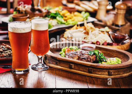 Hot steak with tomatoes, salad, zucchini, greens and sauce on a wooden board. Two glasses of dark beer. Beer table in the restaurant Stock Photo