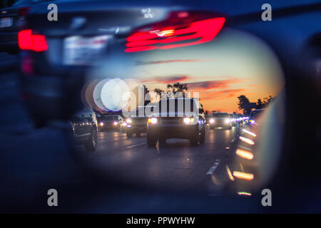 An artistic view of the busy traffic via side mirror at sunset in Los Angeles. Blurred road, headlights and rear lights of other commuters. Stock Photo