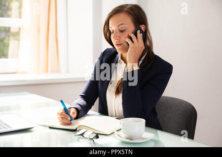 Businesswoman Talking On Cellphone While Writing Schedule In Diary Stock Photo