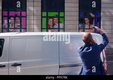 A man takes a picture with one mobile phone while talking into another in front of smiling Superdrug models, in the City of London, the capital's financial heart, on 25th September 2018, in London, England. Stock Photo