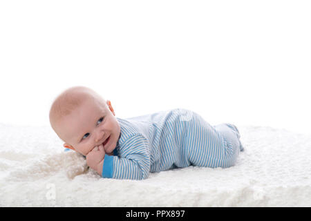 A 2 month old baby boy lying on his stomach on a white blanket. He is wearing pajamas, has his fingers in his mouth and has a mischievous grin. Shot i Stock Photo
