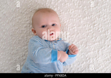 A 2 month old baby boy lying on his back on a white blanket. He is wearing blue and white striped pajamas and is looking at the camera. Stock Photo