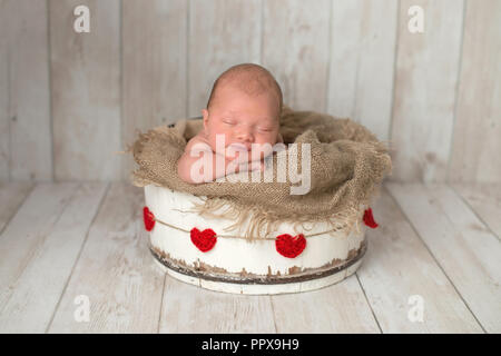 Ten day old newborn baby boy sleeping in a white, wooden bucket with a red, heart garland. Shot in the studio on a light wood background. Stock Photo