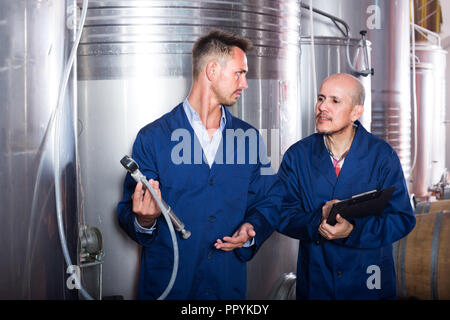 Two  male winery workers  in uniform working and taking notes in secondary fermentation section Stock Photo