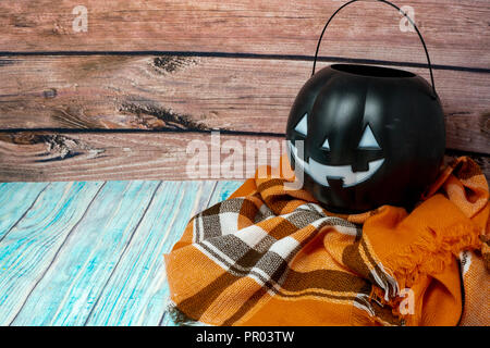 Black jack-o-lantern trick or treat candy bucket pail sitting on a cozy orange plaid blanket. Wood background. Concept for Halloween and fall Stock Photo