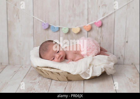 Portrait of a 2 week old newborn baby girl wearing frilly, pink bloomers. She is lying in a wooden bowl and there is a heart garland in the background Stock Photo