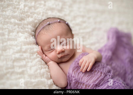 Sleeping, two week old newborn baby girl covered in a lavender purple blanket and wearing a pearl headband. Stock Photo