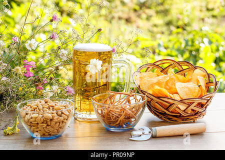 Glass mug of beer on wooden table with potato chips in wicker basket, peanuts and dried squid in bowls on natural green blurred background Stock Photo