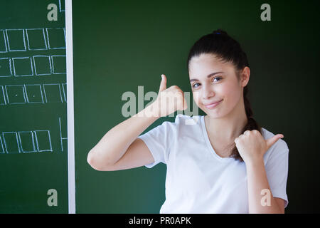 Schoolgirl showing thumbs up against blackboard Stock Photo