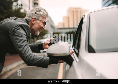 Smiling businessman holding phone and talking with taxi driver on city street. Male traveler communicating with cab driver. Stock Photo